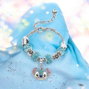 K&ouml;nnte beinhalten: Silbernes Charm-Armband mit blauen und klaren Perlen sowie einem Anh&auml;nger eines Zeichentrickcharakters mit rosa Ohren und blauen Augen. Das Armband liegt auf einem hellblauen Stoff.