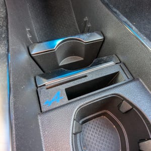 Op de afbeelding: Een zwart plastic opbergvak voor de middenconsole van een auto met een blauw A110-logo, een rechthoekige gleuf en een ronde bekerhouder met een getextureerde bodem. De unit bevat een schuifdeksel met een gebogen uitsparing.
