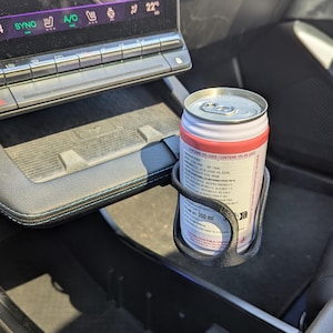 Op de afbeelding: Een zwarte 3D-geprinte bekerhouder bevestigd aan de middenconsole van een voertuig, met daarin een aluminium blikje van 350 ml met een roze en wit etiket. Het blikje bevat voedingsinformatie en de tekst Contains 10% Juice.