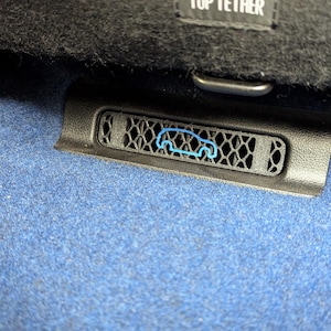 Könnte beinhalten: Eine schwarze Kunststoff-Lüftungsabdeckung für den Fahrzeuginnenraum mit einem blauen Umriss-Symbol eines Autos. Die Lüftung hat ein geometrisches Wabenmuster und befindet sich auf einem blauen Teppichboden unter einer Blende mit TOP TETHER.