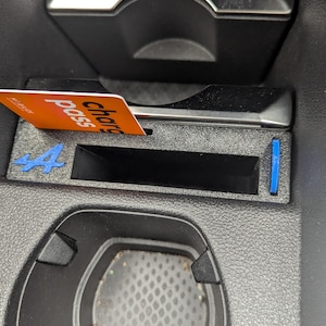 Op de afbeelding: Een zwarte kunststof middenconsole-inzet voor in de auto met een rechthoekig opbergvak, een blauw Alpine-logo aan de linkerkant en een klein blauw verticaal accent aan de rechterkant. Een oranje kaart met de tekst charge pass zit in de gleuf.