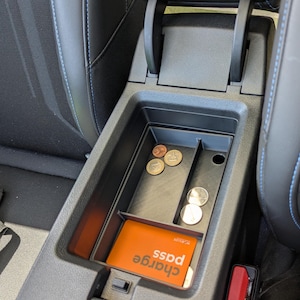 Peut inclure: Un plateau organisateur de console centrale de voiture en plastique noir avec des compartiments divisés. Le plateau contient plusieurs pièces métalliques et une carte rectangulaire orange avec le texte blanc "charge pass" imprimé à sa surface.