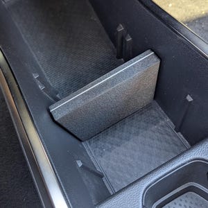 May include: A black plastic center console organizer divider insert with a textured surface. The rectangular divider is positioned vertically within the deep storage compartment of a vehicle, creating a separate section for organized storage.