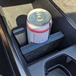 Op de afbeelding: Een zwarte schuimrubberen bekerhouder-inzetstuk met een dunne blauwe horizontale streep, geplaatst in de middenconsole van een voertuig. Een zilveren en rode aluminium drankblikje zit stevig in het schuimrubberen inzetstuk met uitsparing.
