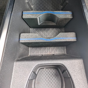 Peut inclure: Console de voiture noire avec deux porte-gobelets et un petit espace de rangement. Les porte-gobelets ont un liseré bleu. La console est en plastique texturé.