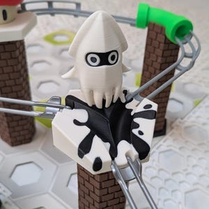 Puede incluir: Un componente de circuito de canicas impreso en 3D que presenta una figura blanca con forma de calamar con ojos negros y un diseño de salpicaduras de tinta negra. El componente es parte de un circuito de canicas.