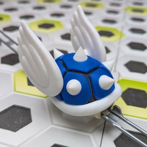 Peut inclure: Une coquille ailée à pointes imprimée en 3D bleue et blanche. La coquille est bleue avec des pointes blanches et des ailes blanches. La coquille est posée sur une surface blanche et grise.