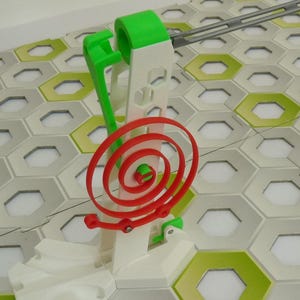 Puede incluir: Primer plano de un componente colorido de una pista de canicas de juguete. La estructura es principalmente blanca con detalles en verde y rojo. La pista está sobre una base de baldosas hexagonales con patrones verdes y grises.