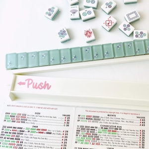 May include: A Mahjong set with white tiles featuring various symbols and a light green tile holder. The set includes a white tray with "Push" and "Play" labels, and a reference sheet with game rules.