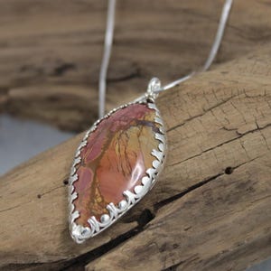 May include: A silver pendant necklace featuring a teardrop-shaped stone with a unique pattern of red, orange, and yellow hues. The stone is encased in a decorative silver setting and hangs from a delicate silver chain.
