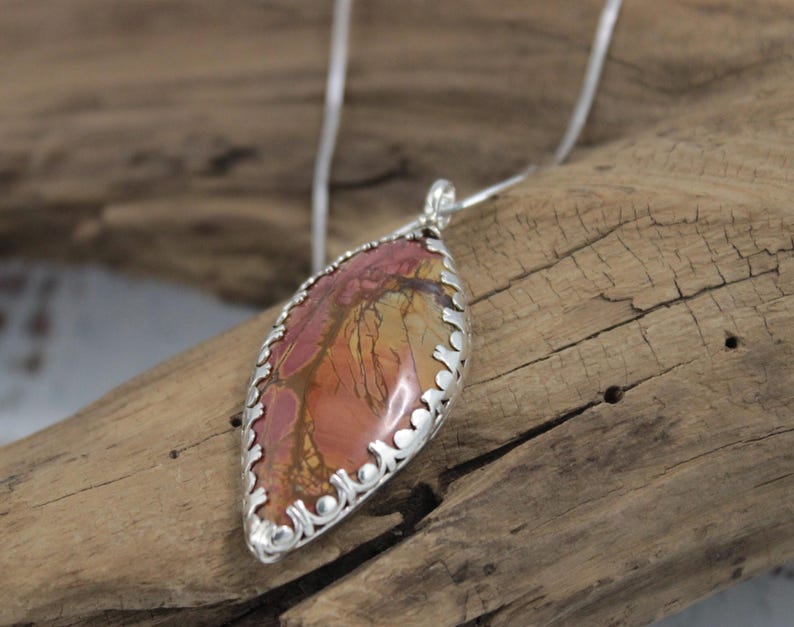 May include: A silver pendant necklace featuring a teardrop-shaped stone with a unique pattern of red, orange, and yellow hues. The stone is encased in a decorative silver setting and hangs from a delicate silver chain.
