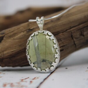 May include: Oval pendant necklace featuring a green and gray stone set in a decorative silver-tone frame. The pendant hangs from a delicate silver chain, showcasing a natural, earthy aesthetic. The stone has a unique pattern.