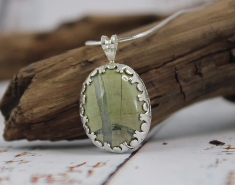 May include: Oval pendant necklace featuring a green and gray stone set in a decorative silver-tone frame. The pendant hangs from a delicate silver chain, showcasing a natural, earthy aesthetic. The stone has a unique pattern.