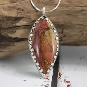 May include: A pendant necklace featuring a large, oval-shaped stone with a silver-colored, scalloped bezel. The stone displays a mix of red, brown, and yellow hues. The necklace chain is a thin, silver-colored metal.