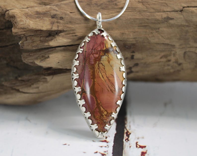 May include: A pendant necklace featuring a large, oval-shaped stone with a silver-colored, scalloped bezel. The stone displays a mix of red, brown, and yellow hues. The necklace chain is a thin, silver-colored metal.