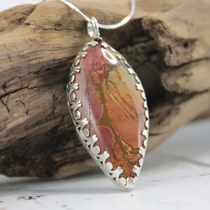 May include: A silver pendant necklace featuring a large, oval-shaped stone with a mix of red, orange, and yellow hues. The stone is set in a decorative silver frame. The necklace is displayed on a piece of driftwood.