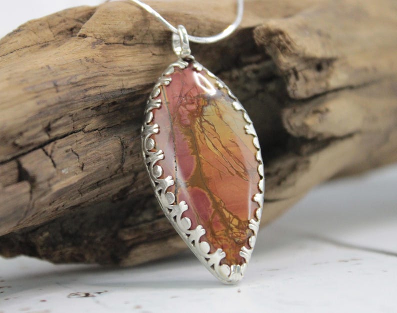 May include: A silver pendant necklace featuring a large, oval-shaped stone with a mix of red, orange, and yellow hues. The stone is set in a decorative silver frame. The necklace is displayed on a piece of driftwood.