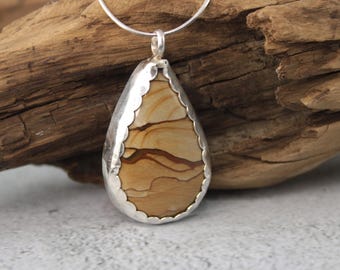 May include: A teardrop-shaped pendant featuring a tan and brown banded stone set in a scalloped silver bezel. The pendant hangs from a thin silver chain, showcasing natural horizontal layers of earth-toned mineral patterns within the stone.