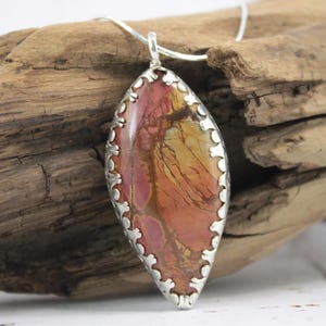 May include: A silver pendant necklace featuring a large, oval-shaped stone with a unique pattern of red, orange, and yellow hues. The stone is set in a decorative silver frame and hangs from a delicate silver chain. The pendant is displayed against a rustic wooden background.