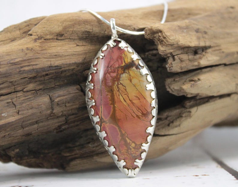 May include: A silver pendant necklace featuring a large, oval-shaped stone with a unique pattern of red, orange, and yellow hues. The stone is set in a decorative silver frame and hangs from a delicate silver chain. The pendant is displayed against a rustic wooden background.