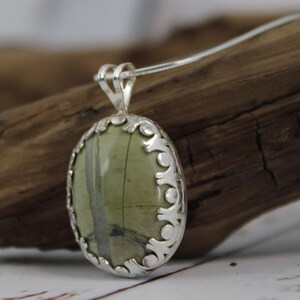 May include: Oval pendant necklace featuring a green stone with gray veining, set in a decorative silver-toned frame. The pendant hangs from a delicate silver chain, showcasing a natural gemstone design.
