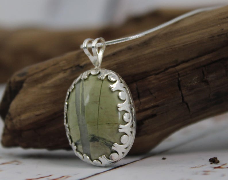 May include: Oval pendant necklace featuring a green stone with gray veining, set in a decorative silver-toned frame. The pendant hangs from a delicate silver chain, showcasing a natural gemstone design.