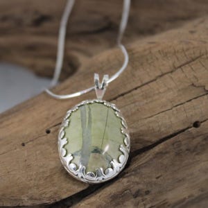 May include: Oval pendant necklace featuring a green and beige stone set in a decorative silver-tone frame. The pendant hangs from a delicate silver chain, creating an elegant and natural look. The necklace is displayed on a wooden surface.