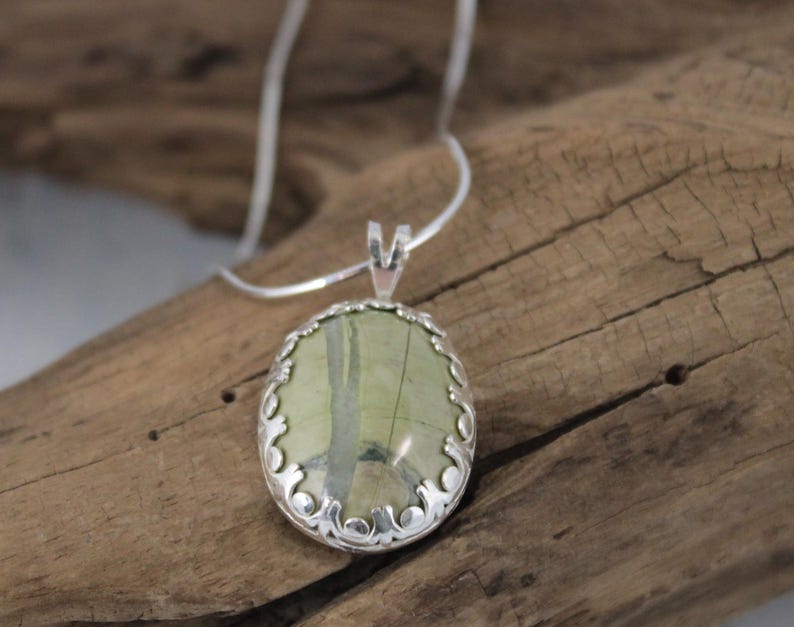 May include: Oval pendant necklace featuring a green and beige stone set in a decorative silver-tone frame. The pendant hangs from a delicate silver chain, creating an elegant and natural look. The necklace is displayed on a wooden surface.