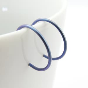 May include: A pair of small, round hoop earrings made of blue titanium. The earrings are smooth and have a subtle iridescent sheen.