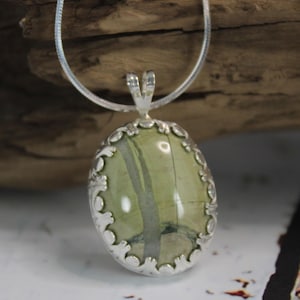 May include: Oval pendant with a green stone and silver-colored filigree setting. The stone has a natural pattern of green and gray. The pendant hangs from a silver-colored chain, creating a classic jewelry piece.
