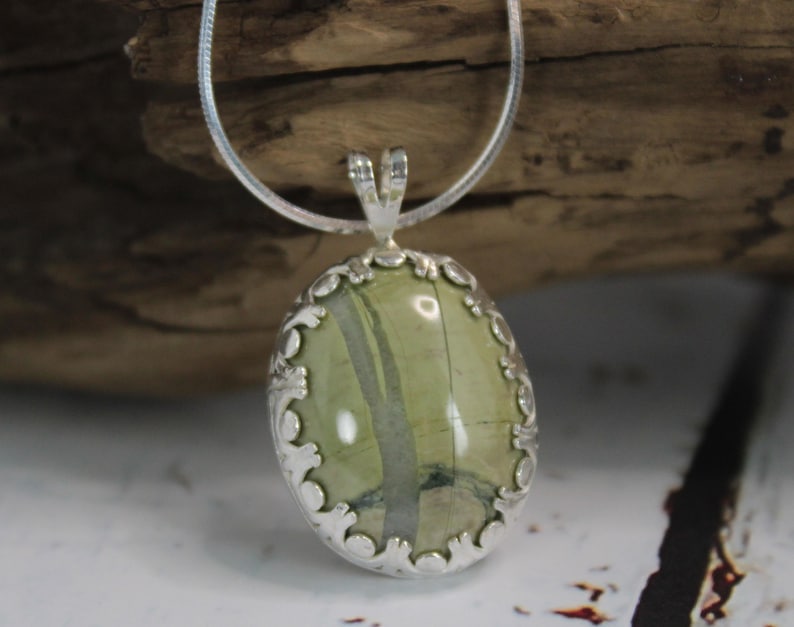 May include: Oval pendant with a green stone and silver-colored filigree setting. The stone has a natural pattern of green and gray. The pendant hangs from a silver-colored chain, creating a classic jewelry piece.
