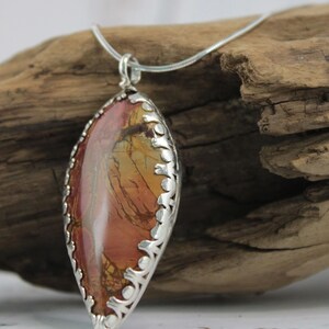May include: A pendant necklace featuring a large, oval-shaped stone with a silver filigree setting. The stone displays a blend of red, orange, and brown hues. The necklace is displayed on a piece of driftwood.