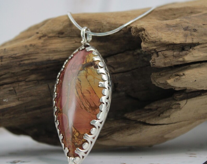May include: A pendant necklace featuring a large, oval-shaped stone with a silver filigree setting. The stone displays a blend of red, orange, and brown hues. The necklace is displayed on a piece of driftwood.