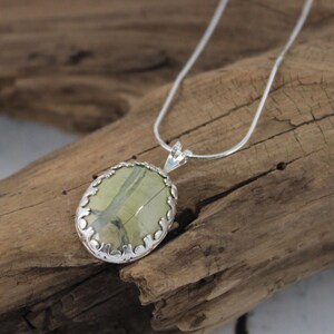 May include: Oval pendant necklace featuring a light green stone with darker green veining, set in a silver-colored filigree frame. The pendant hangs from a delicate silver chain, displayed on a piece of driftwood.