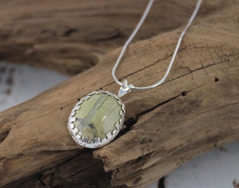 May include: Oval pendant necklace featuring a light green stone with darker green veining, set in a silver-colored filigree frame. The pendant hangs from a delicate silver chain, displayed on a piece of driftwood.