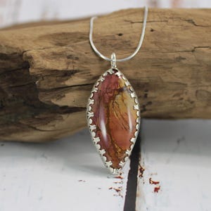 May include: A silver pendant necklace featuring an oval-shaped stone with a mix of red, orange, and yellow hues. The stone is encased in a decorative silver setting and hangs from a delicate silver chain. The pendant rests on a piece of driftwood.