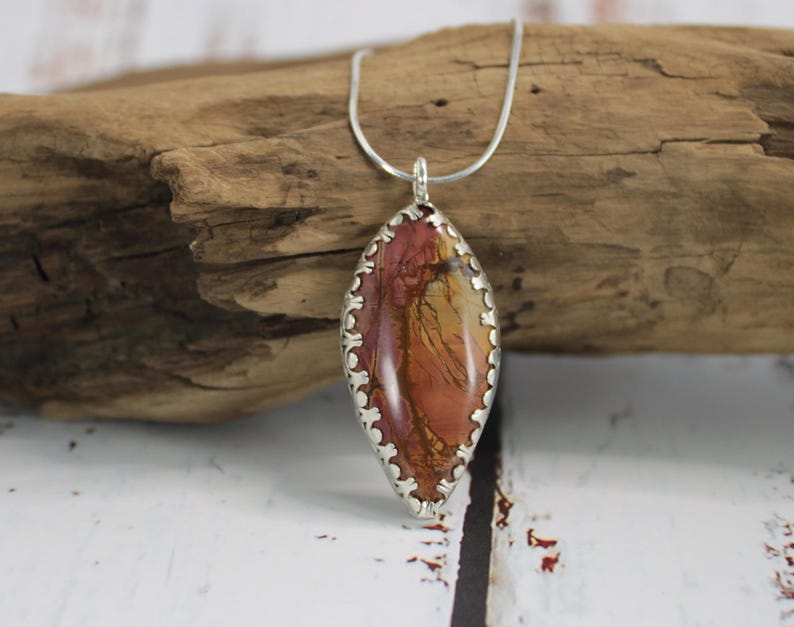 May include: A silver pendant necklace featuring an oval-shaped stone with a mix of red, orange, and yellow hues. The stone is encased in a decorative silver setting and hangs from a delicate silver chain. The pendant rests on a piece of driftwood.