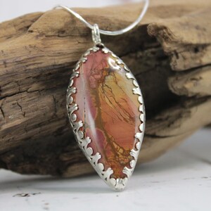 May include: A pendant necklace featuring a large, oval-shaped stone with a silver-colored filigree setting. The stone displays a blend of red, orange, and yellow hues. The necklace is displayed on a piece of driftwood.