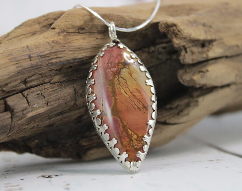 May include: A pendant necklace featuring a large, oval-shaped stone with a silver-colored filigree setting. The stone displays a blend of red, orange, and yellow hues. The necklace is displayed on a piece of driftwood.