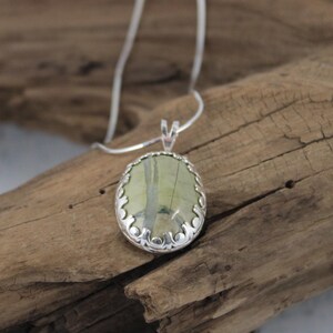 May include: A silver pendant necklace featuring an oval-shaped, light green stone with darker green veining. The stone is set in a decorative silver bezel and hangs from a delicate silver chain. The necklace is displayed on a piece of weathered wood.