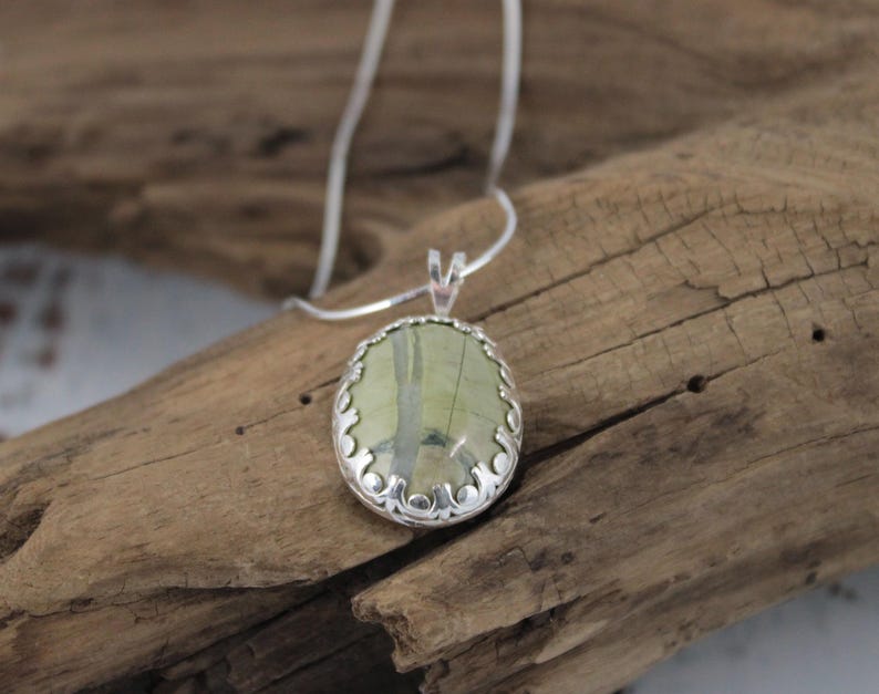 May include: A silver pendant necklace featuring an oval-shaped, light green stone with darker green veining. The stone is set in a decorative silver bezel and hangs from a delicate silver chain. The necklace is displayed on a piece of weathered wood.