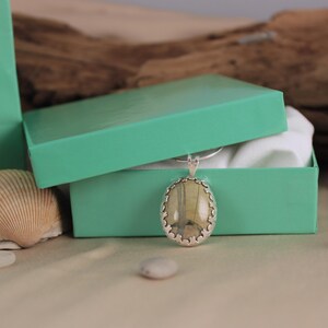 May include: A silver pendant with an oval, light-colored stone is displayed in an open, turquoise gift box. The pendant has a decorative silver border. A seashell and small white stones are in the foreground.