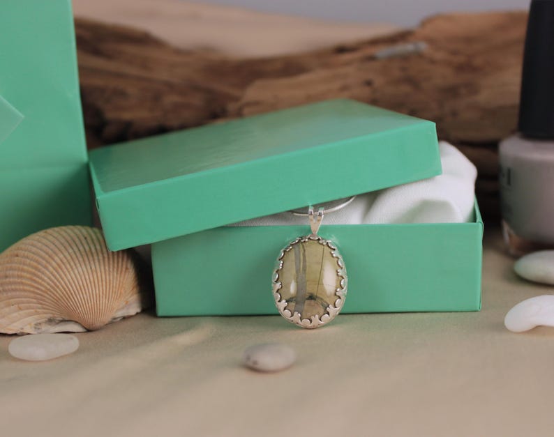 May include: A silver pendant with an oval, light-colored stone is displayed in an open, turquoise gift box. The pendant has a decorative silver border. A seashell and small white stones are in the foreground.