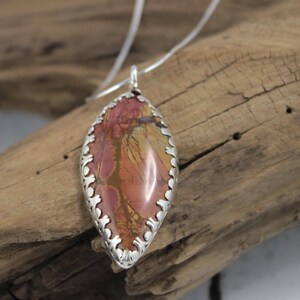 May include: A pendant necklace featuring a teardrop-shaped stone with a silver filigree border. The stone has a mix of red, orange, and brown hues. The necklace is displayed on a wooden surface.