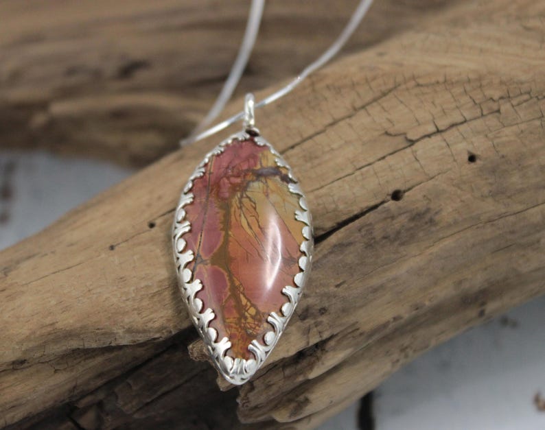 May include: A pendant necklace featuring a teardrop-shaped stone with a silver filigree border. The stone has a mix of red, orange, and brown hues. The necklace is displayed on a wooden surface.