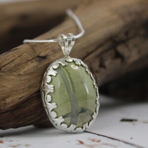 May include: Oval pendant necklace featuring a green stone with dark green veining, set in a decorative silver-toned bezel. The pendant hangs from a delicate silver chain, showcasing a natural, earthy aesthetic. The necklace is a piece of jewelry.