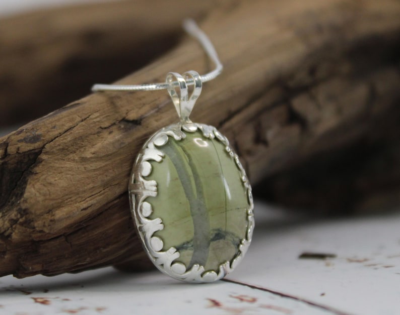 May include: Oval pendant necklace featuring a green stone with dark green veining, set in a decorative silver-toned bezel. The pendant hangs from a delicate silver chain, showcasing a natural, earthy aesthetic. The necklace is a piece of jewelry.