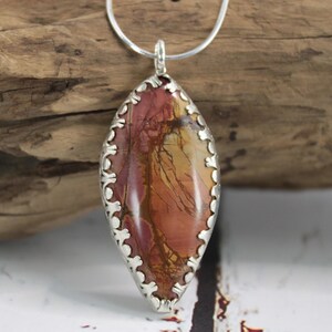 May include: A silver pendant necklace featuring a large, oval-shaped stone with a natural, earthy pattern in shades of red, orange, and yellow. The stone is set in a decorative silver bezel and hangs from a delicate silver chain. The pendant is approximately 2 inches long.