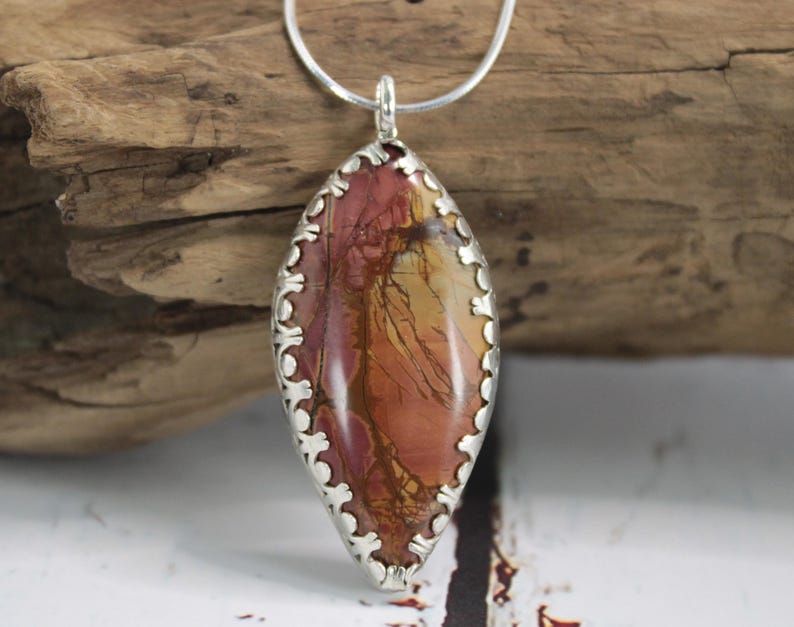 May include: A silver pendant necklace featuring a large, oval-shaped stone with a natural, earthy pattern in shades of red, orange, and yellow. The stone is set in a decorative silver bezel and hangs from a delicate silver chain. The pendant is approximately 2 inches long.