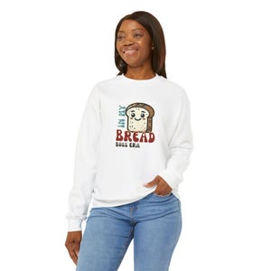 May include: White sweatshirt with a graphic of a smiling bread slice and the text "IN MY BREAD BOSS ERA" in teal and red. The sweatshirt is long-sleeved and made of a soft material.